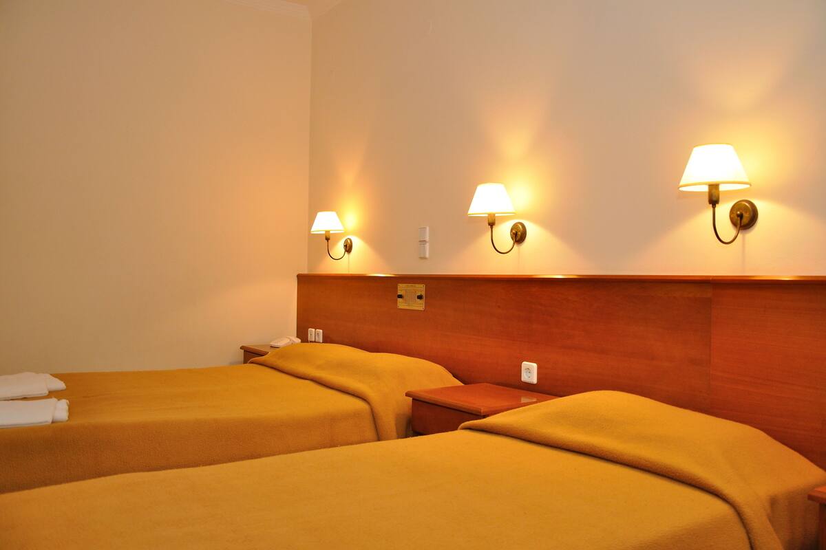 A hotel room with two beds, wooden headboard, wall-mounted lamps, and a small wooden nightstand.