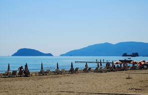 Una playa con filas de sillas de playa y sombrillas, un mar tranquilo y montañas al fondo.