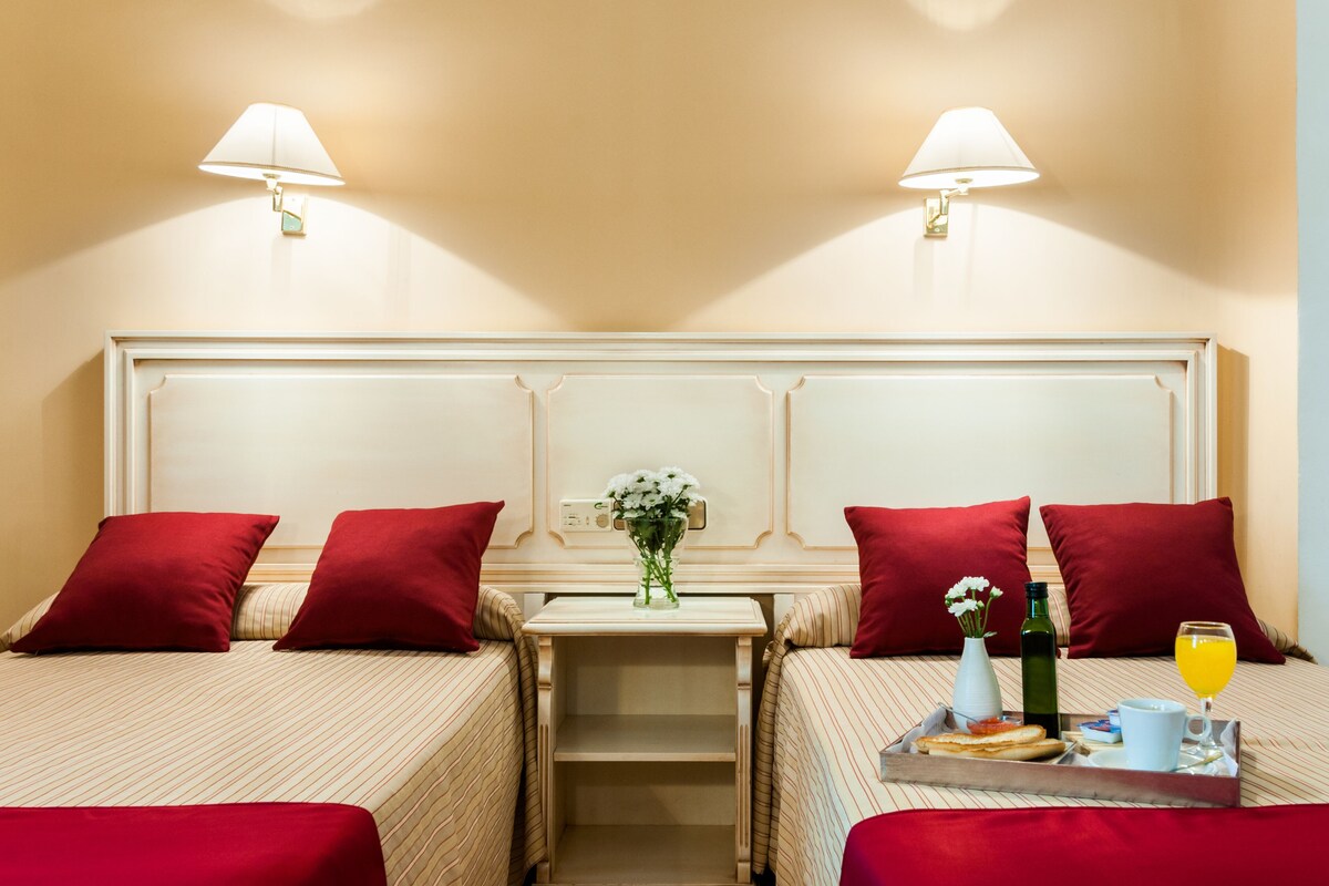 Habitación de hotel con dos camas, cada una con almohada roja, una mesita con un jarrón de flores, una botella y una bandeja con alimentos para el desayuno.