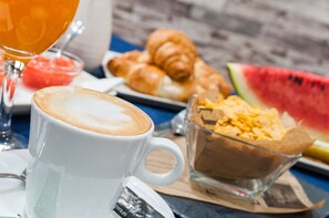 Un desayuno con una taza de café, croissants, un vaso de zumo de naranja y un bol de cereales.