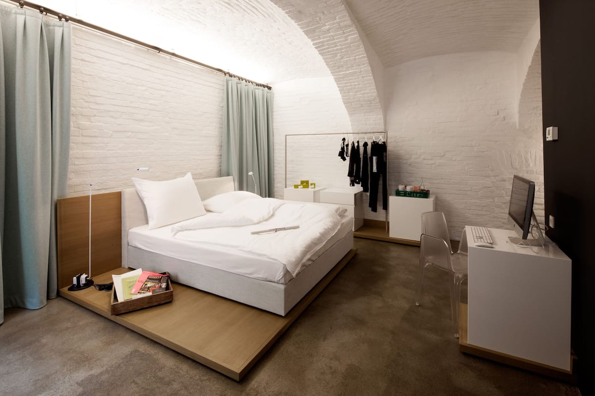 A modern hotel room with a white brick wall, a bed with white linens, a desk with a computer, and a clothing rack.