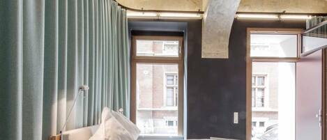 A bedroom with a bed, curtains, a lamp, and a view of buildings through large windows.