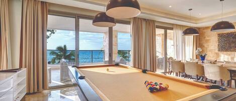 A pool table in a well-lit room with large windows, a dining area, and a view of the ocean.