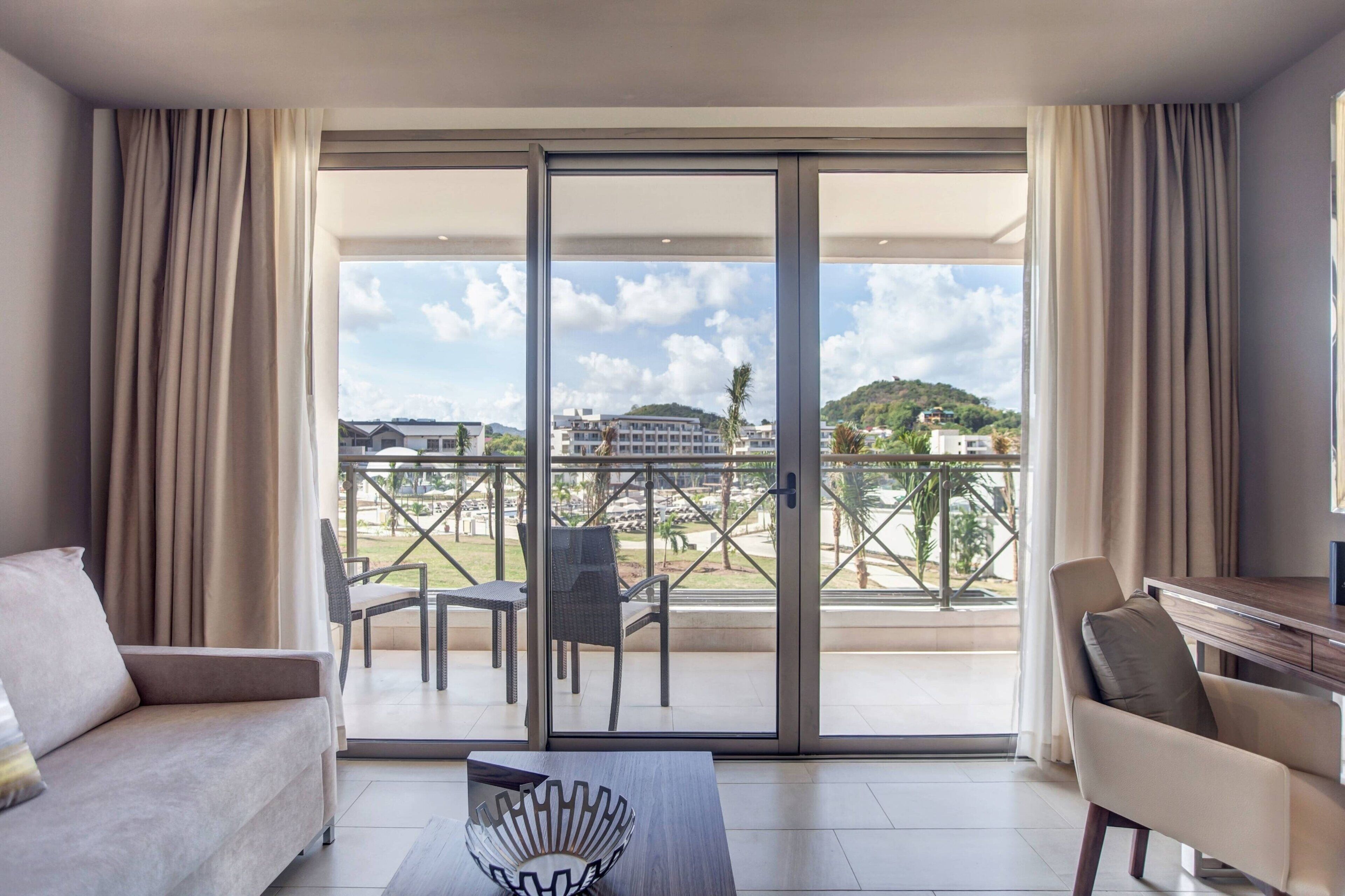 Una habitación de hotel moderna con balcón, sofá, mesa de centro y zona de comedor con sillas.