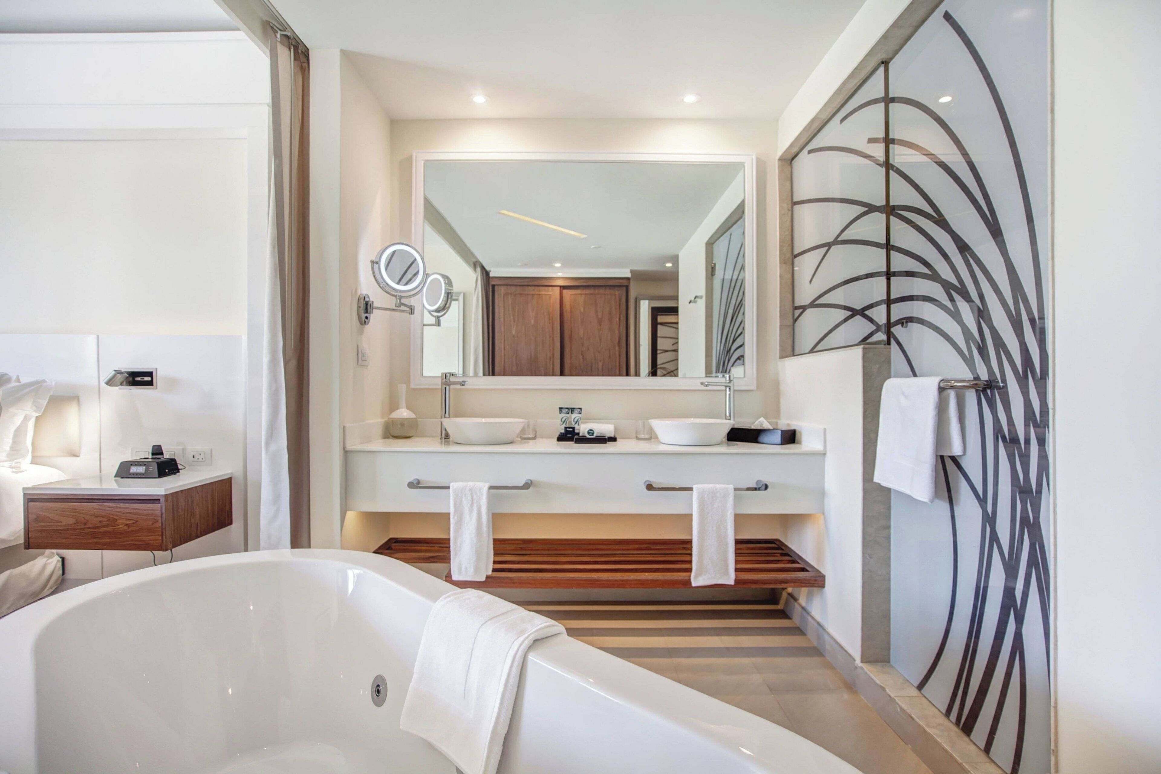 A modern bathroom with a freestanding bathtub, two sinks, a large mirror, and a decorative wall panel.
