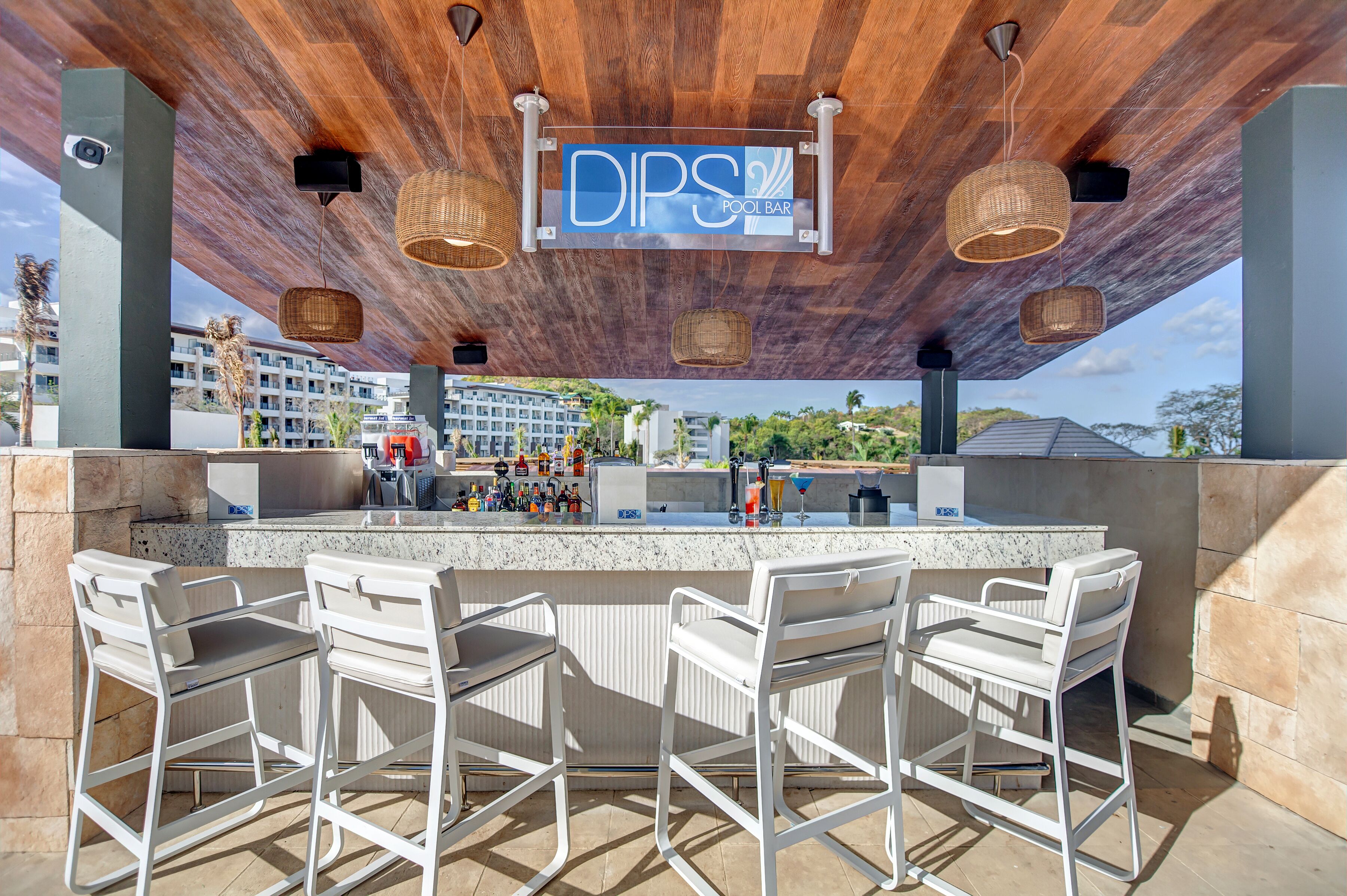 Un espace bar extérieur avec une enseigne indiquant « DPS Pool Bar », doté d’un plafond en bois, de luminaires suspendus en osier et de tabourets de bar blancs.