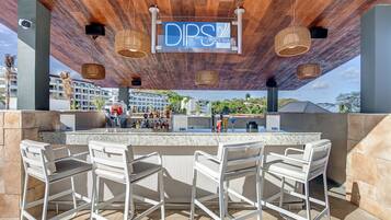 Un espace bar extérieur avec une enseigne indiquant « DPS Pool Bar », doté d’un plafond en bois, de luminaires suspendus en osier et de tabourets de bar blancs.
