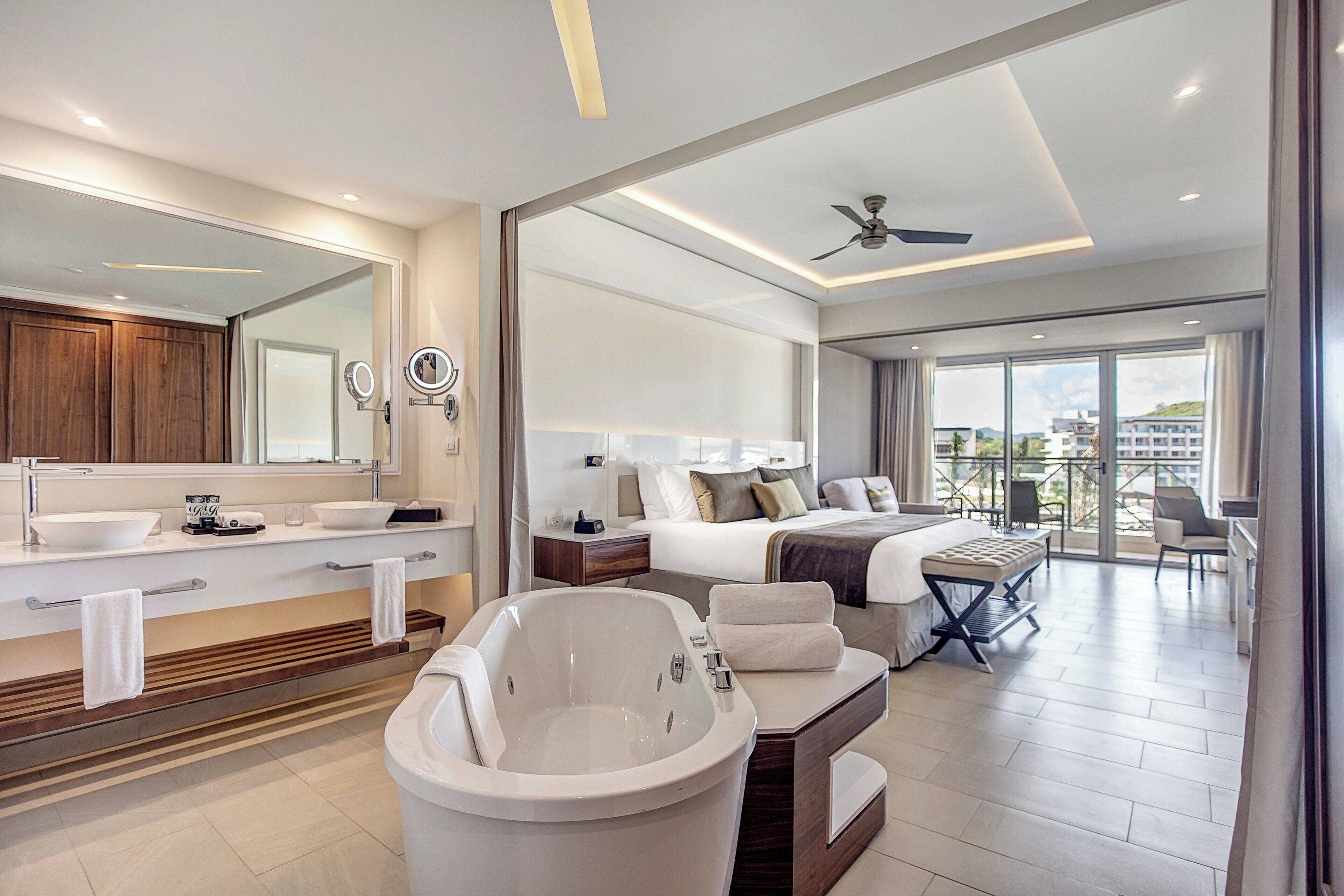 A modern hotel room with a large bathtub, a separate bedroom with a balcony, and a large window offering a city view.