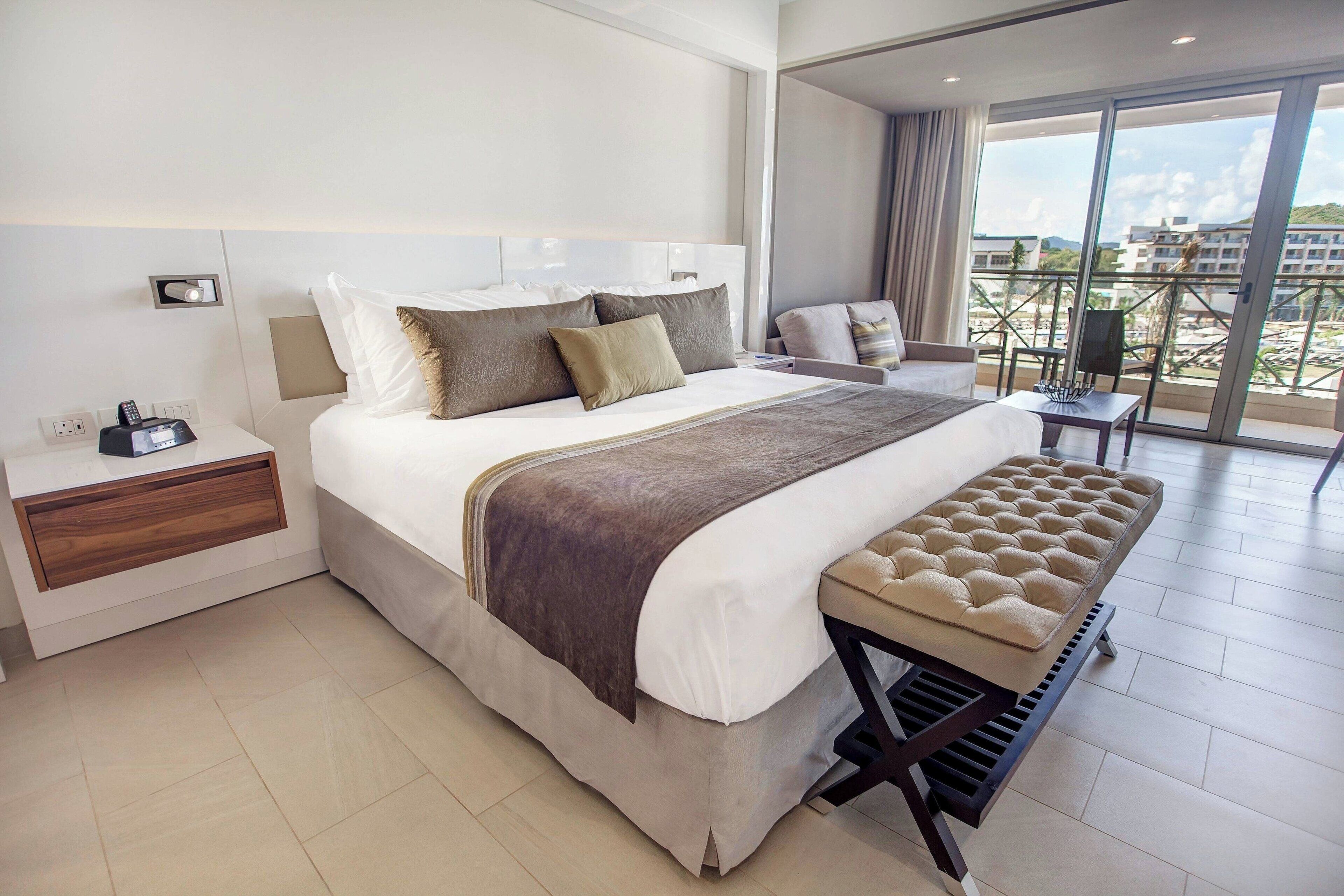 A modern hotel room with a large bed, bedside tables, a bench, and a balcony with a view.