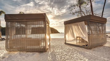Dos cabañas de mimbre en la playa con cortinas blancas, ubicadas en una arena con palmeras y vista al océano.