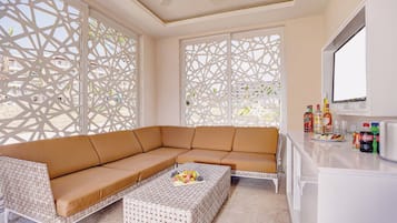 A modern living room with a sectional sofa, a coffee table with fruit, and a kitchen area with a refrigerator and bottles.