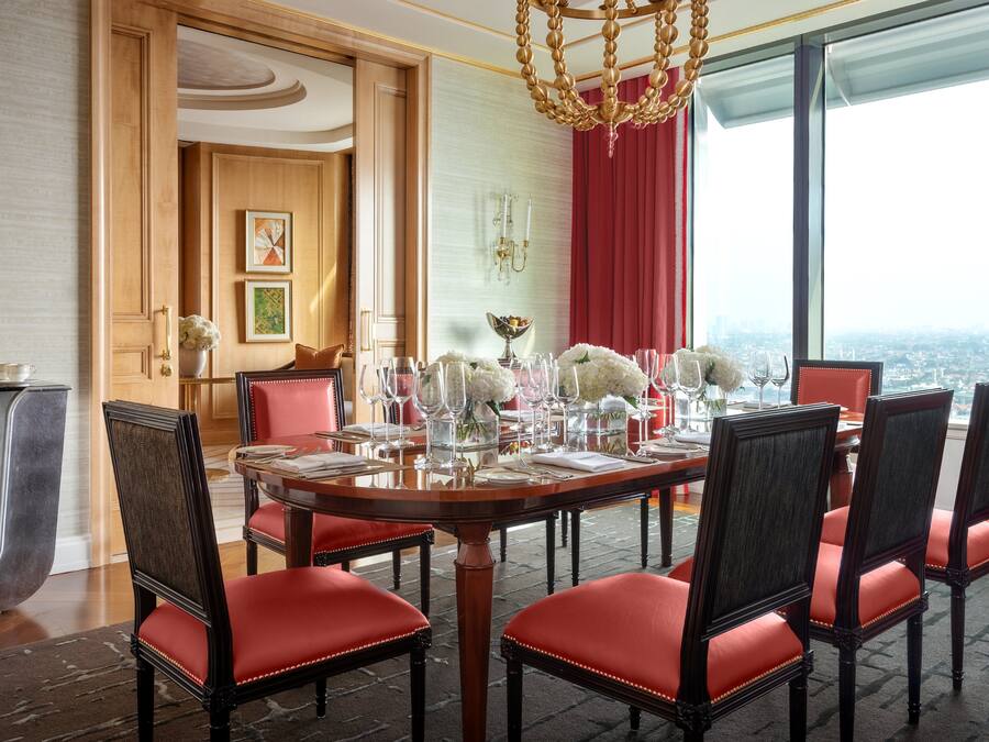 A dining area with a round table set for six, red upholstered chairs, a chandelier, and a city view through large windows.