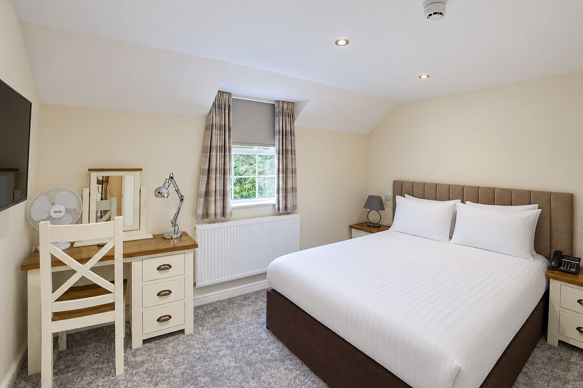 A bedroom with a bed, a desk with a chair, a window with curtains, and a radiator.