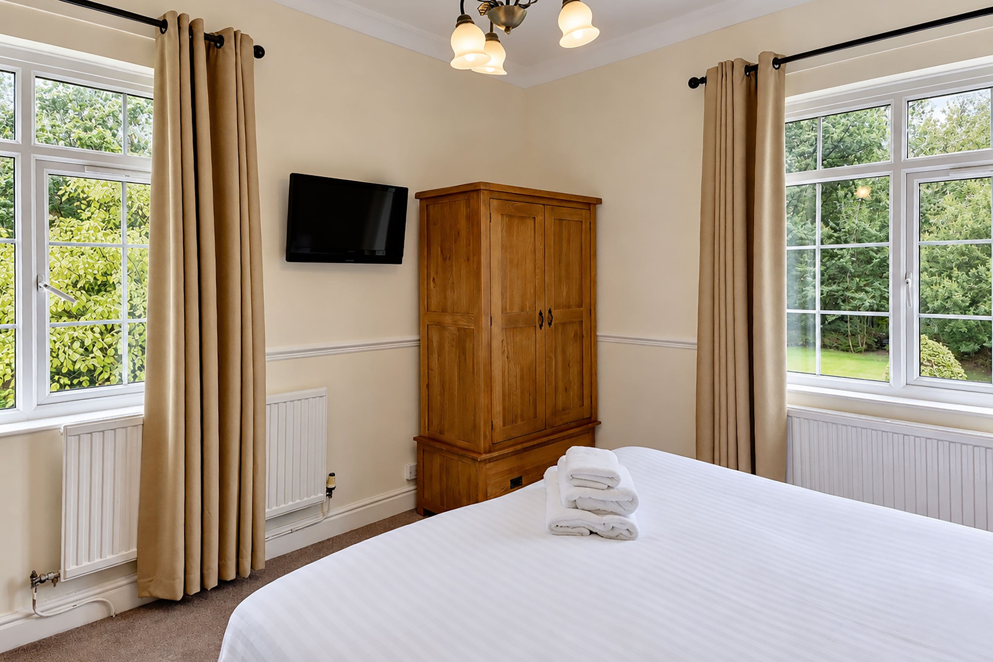 A bedroom with a bed, a wooden wardrobe, a television on the wall, and windows with curtains.