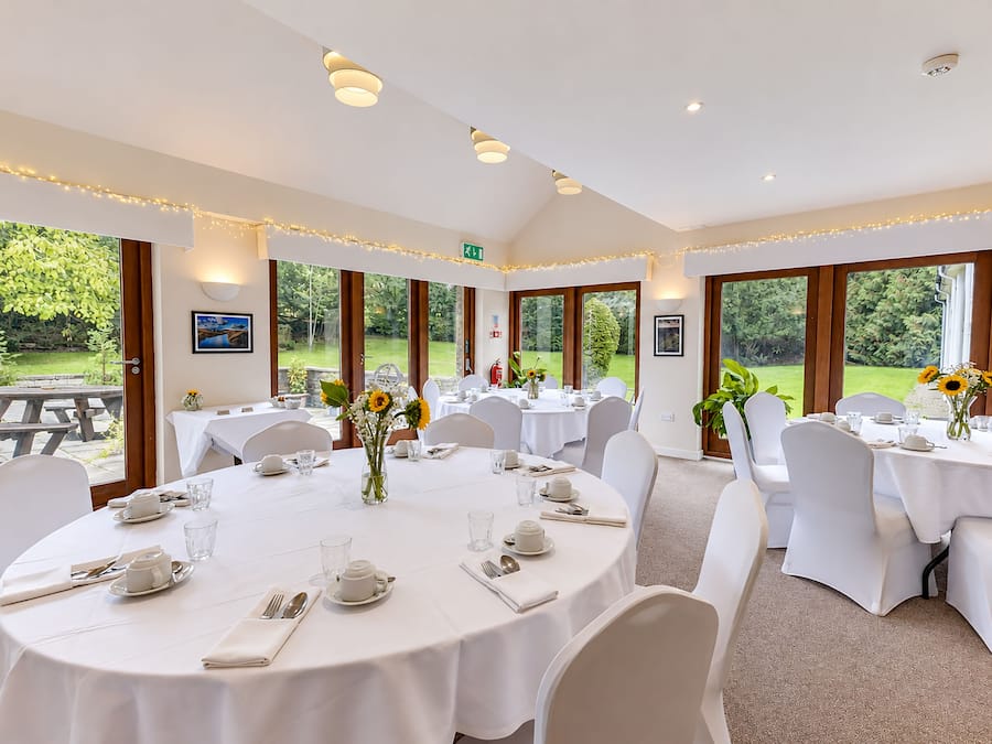 A dining area with round tables, white tablecloths, and chairs, featuring large windows with a view of greenery.