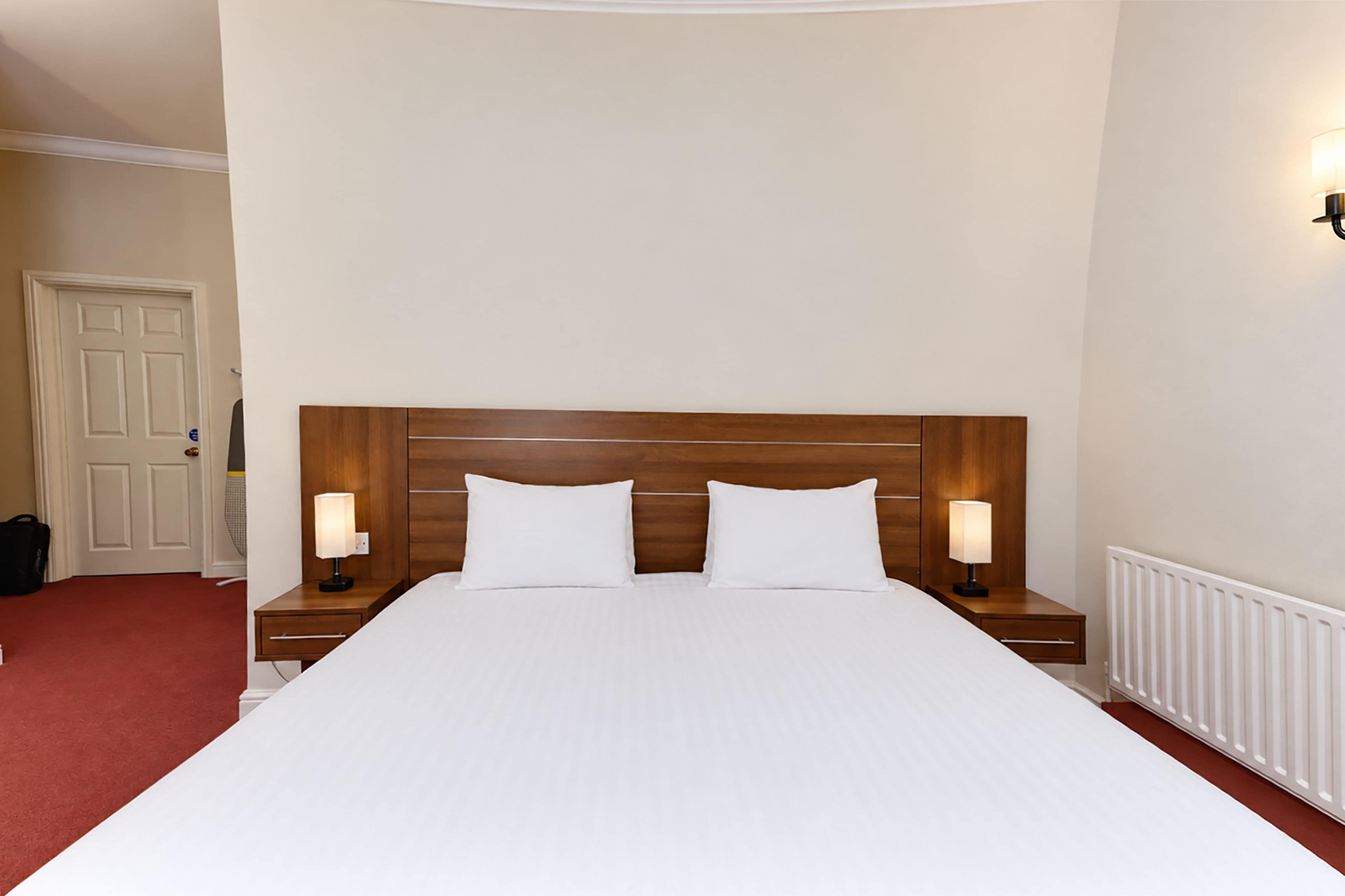 A hotel room with a large bed, two bedside tables with lamps, a wooden headboard, and a white radiator.