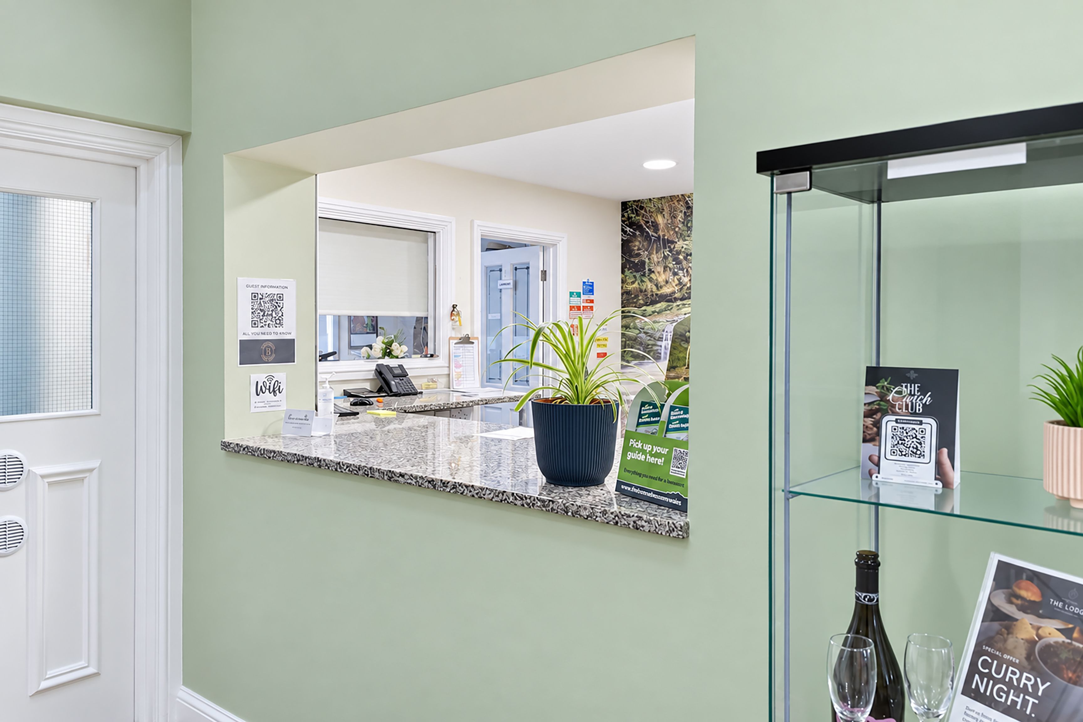 Un espace d’accueil avec un comptoir, un miroir reflétant l’intérieur, une vitrine contenant une bouteille et un verre, et un panneau avec un code QR.