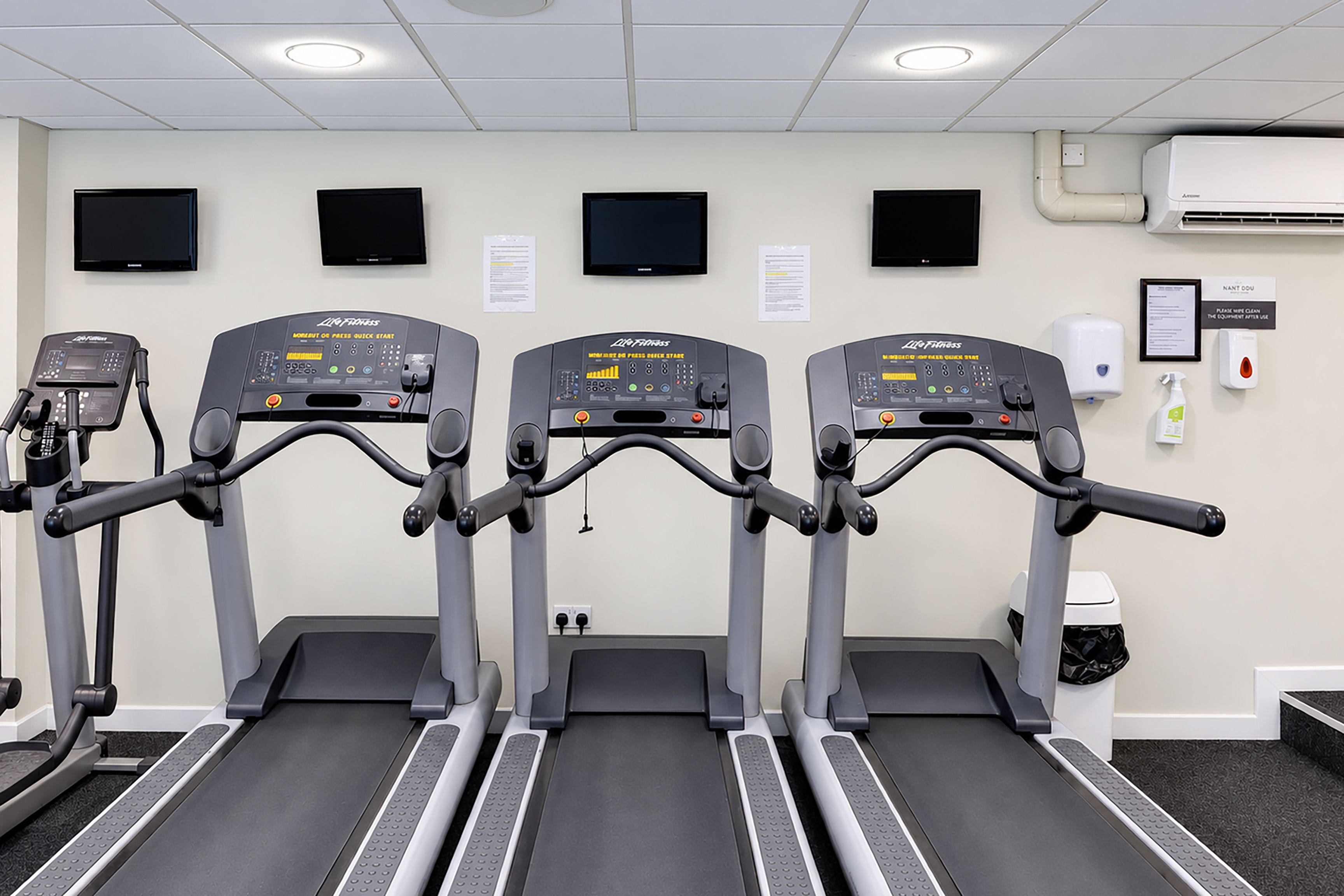 Drei Laufbänder in einem Fitnessstudio mit an der Wand montierten Fernsehern und einem Handdesinfektionsmittelspender.
