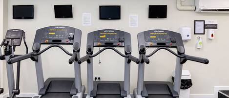 Three treadmills in a gym with wall-mounted televisions and a hand sanitizer dispenser.