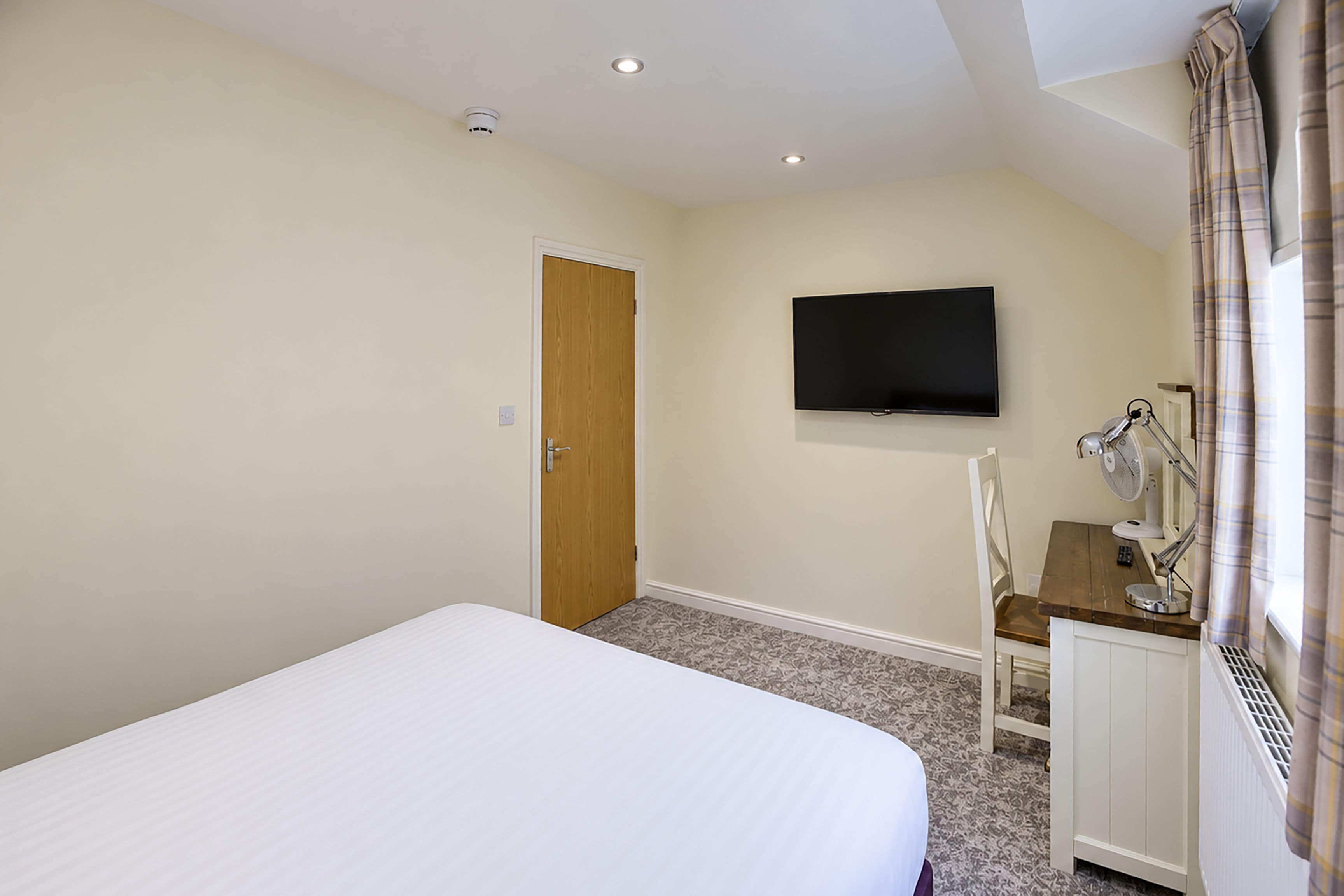 A hotel room with a bed, a television mounted on the wall, a desk with a chair, and a window with curtains.