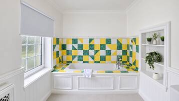 Une salle de bain avec un mur recouvert de carreaux à motifs, une baignoire, une fenêtre avec un store et une étagère ornée de plantes.