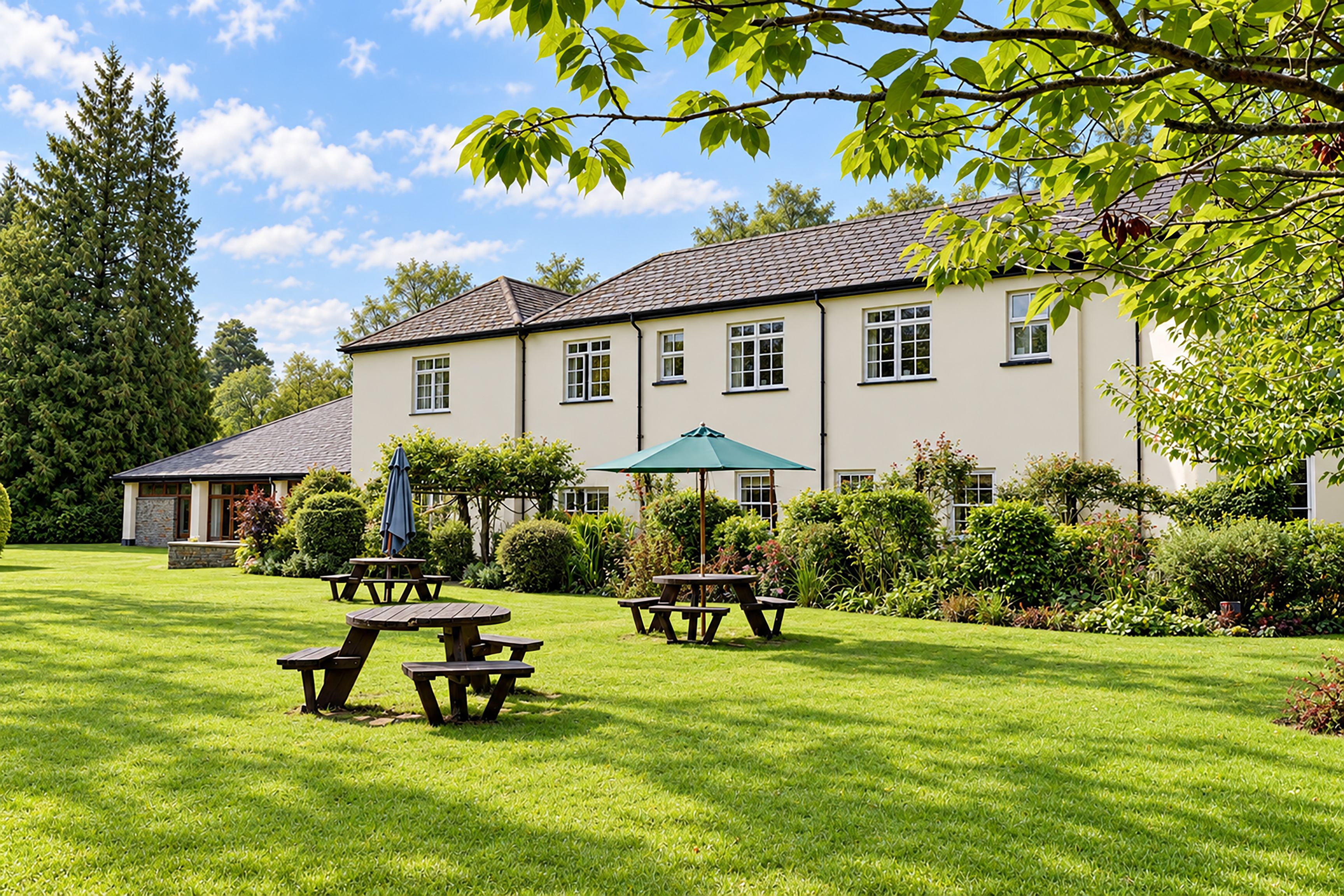 A large building with multiple windows, a lawn with picnic tables, and a tree with green leaves.