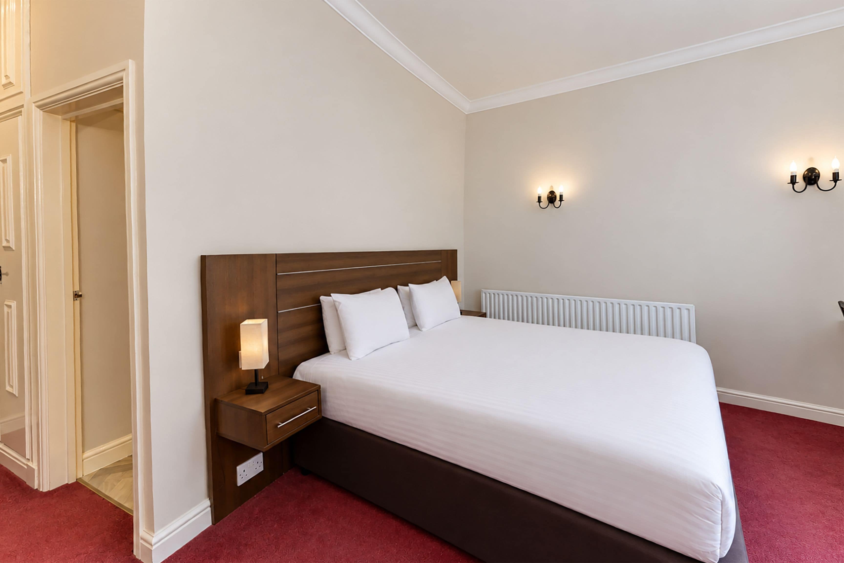 A hotel room with a bed, bedside table, lamp, and wall sconces.