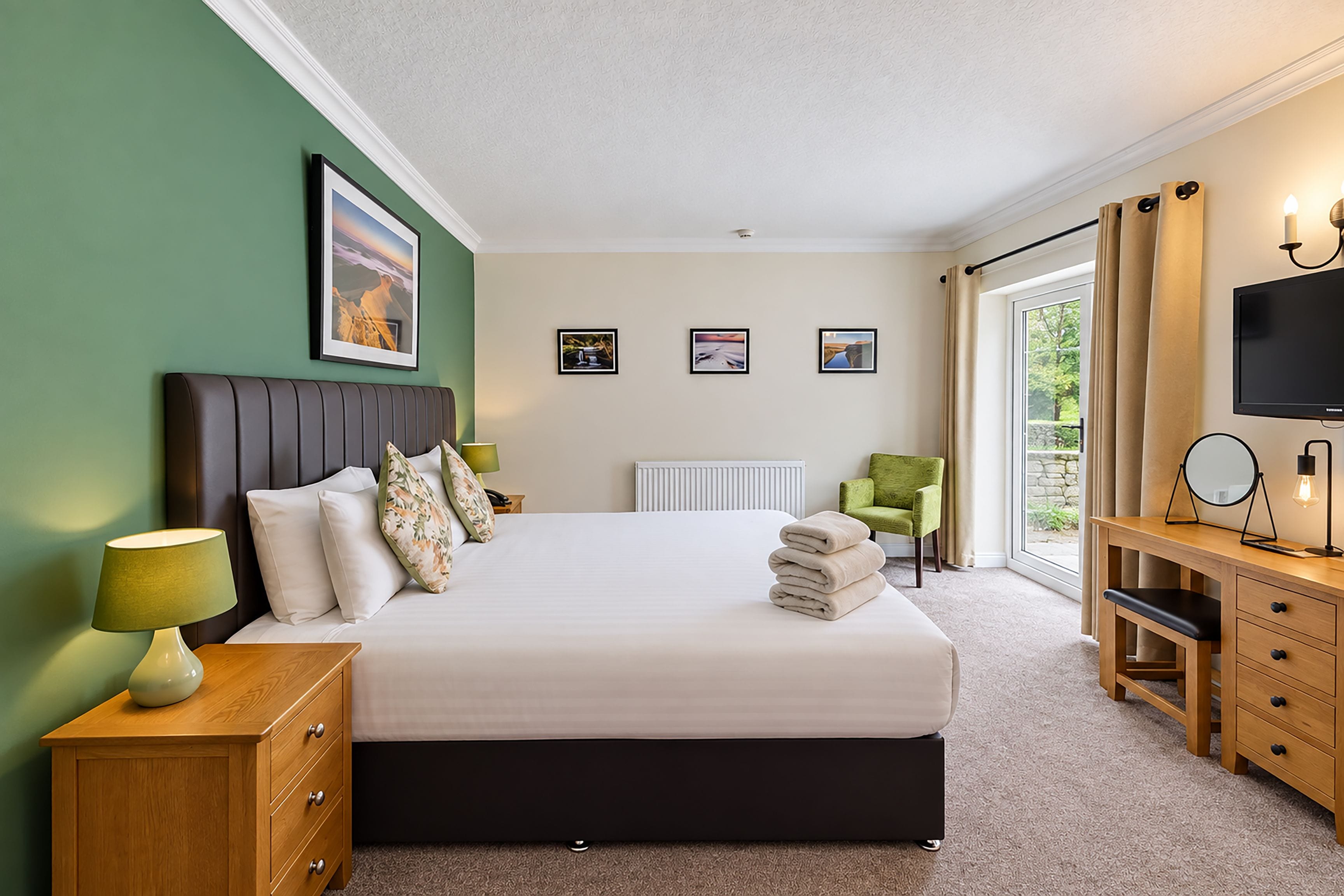 Quarto de hotel com cama, mesas de cabeceira, escrivaninha, cadeira, televisão e quadros nas paredes.