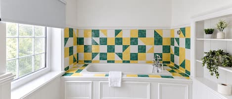 A bathroom with a checkered tile backsplash, a white bathtub, and built-in shelving with plants.