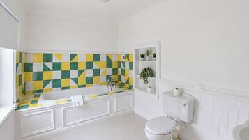 Casa de banho com banheira branca, sanita e lavatório. Tem um painel de azulejos xadrez atrás da banheira.