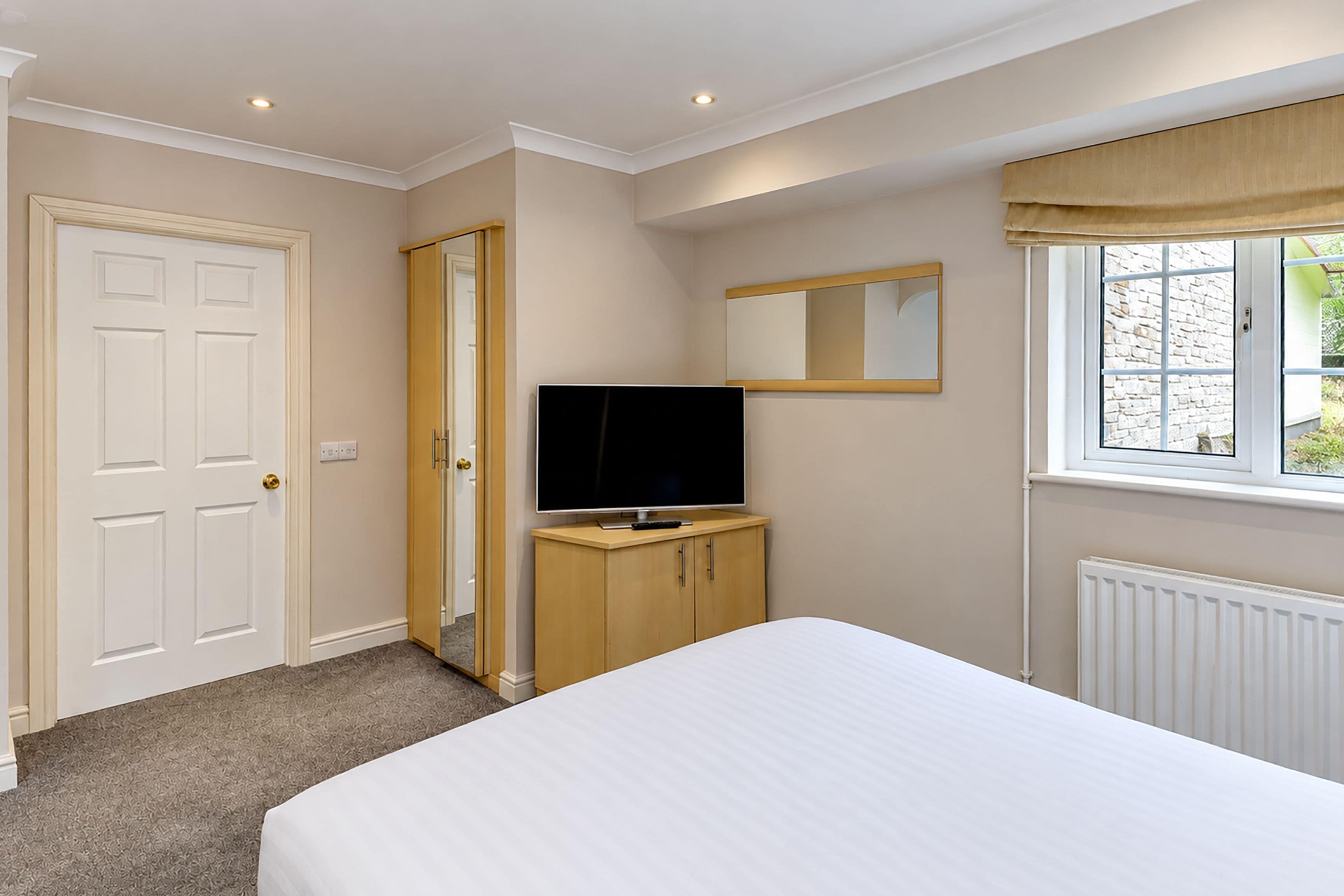 A bedroom with a bed, a television on a wooden cabinet, a mirror, and a window with a view of the outside.