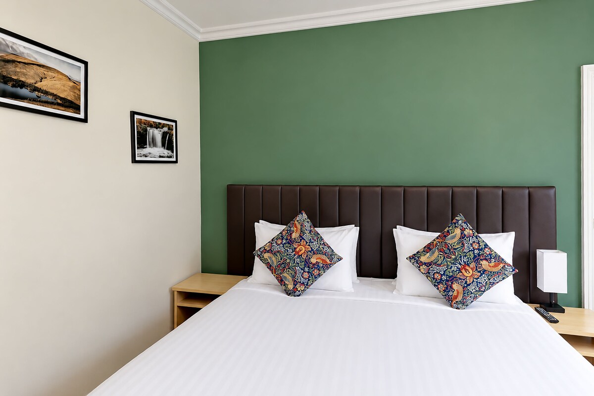 A hotel room with a large bed, two pillows, a nightstand, a bedside table, and a wall-mounted lamp.