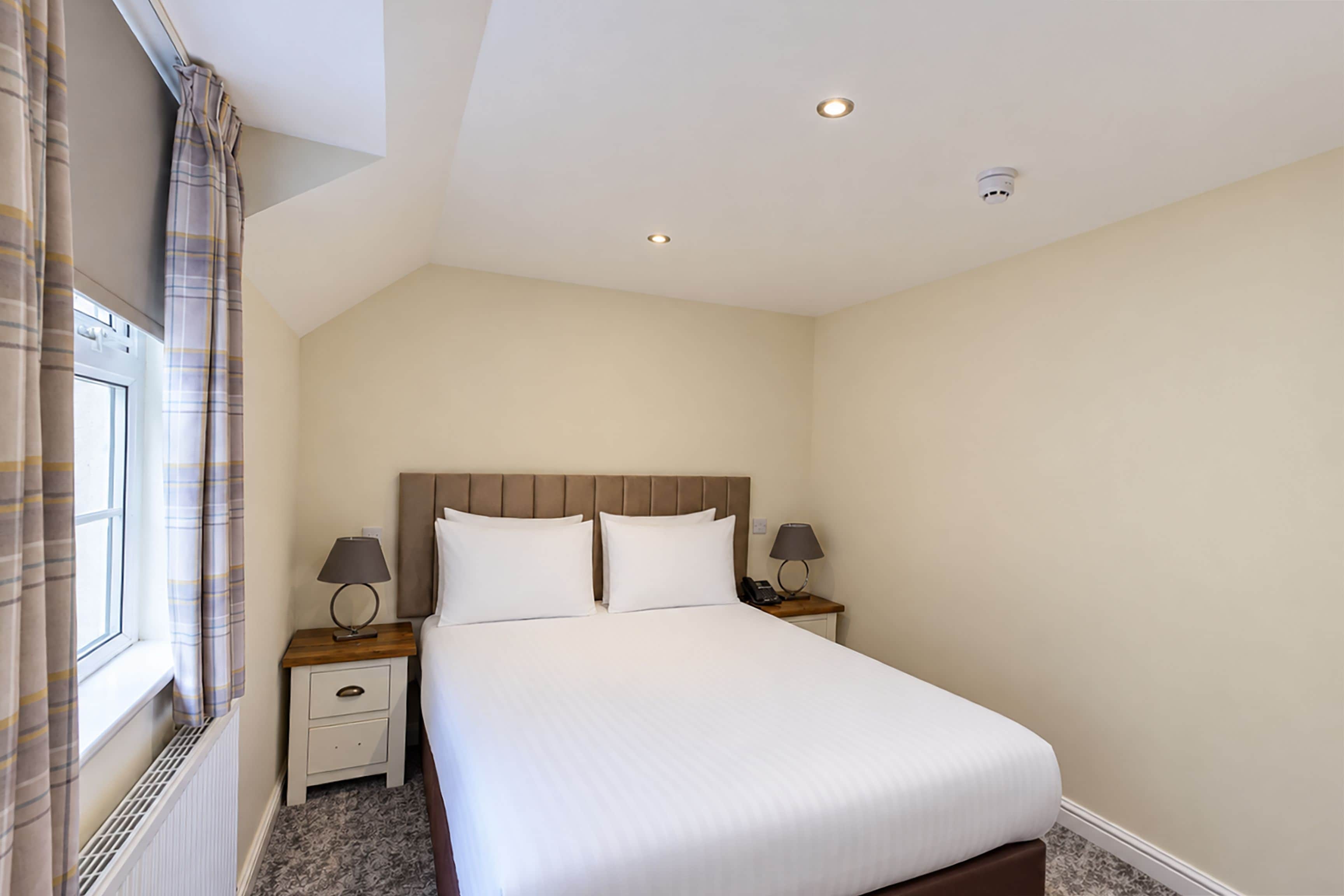 A hotel room with a large bed, two bedside tables with lamps, a window with curtains, and a ceiling with recessed lighting.