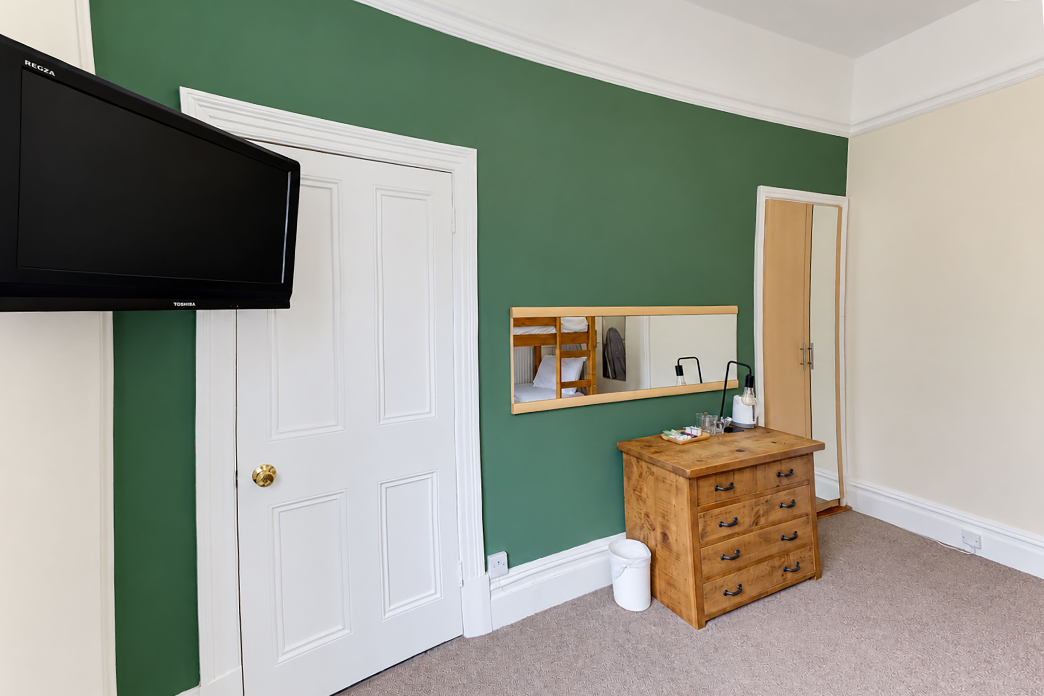 A room with a white door, a mounted television, a wooden dresser, and a mirror on the wall.