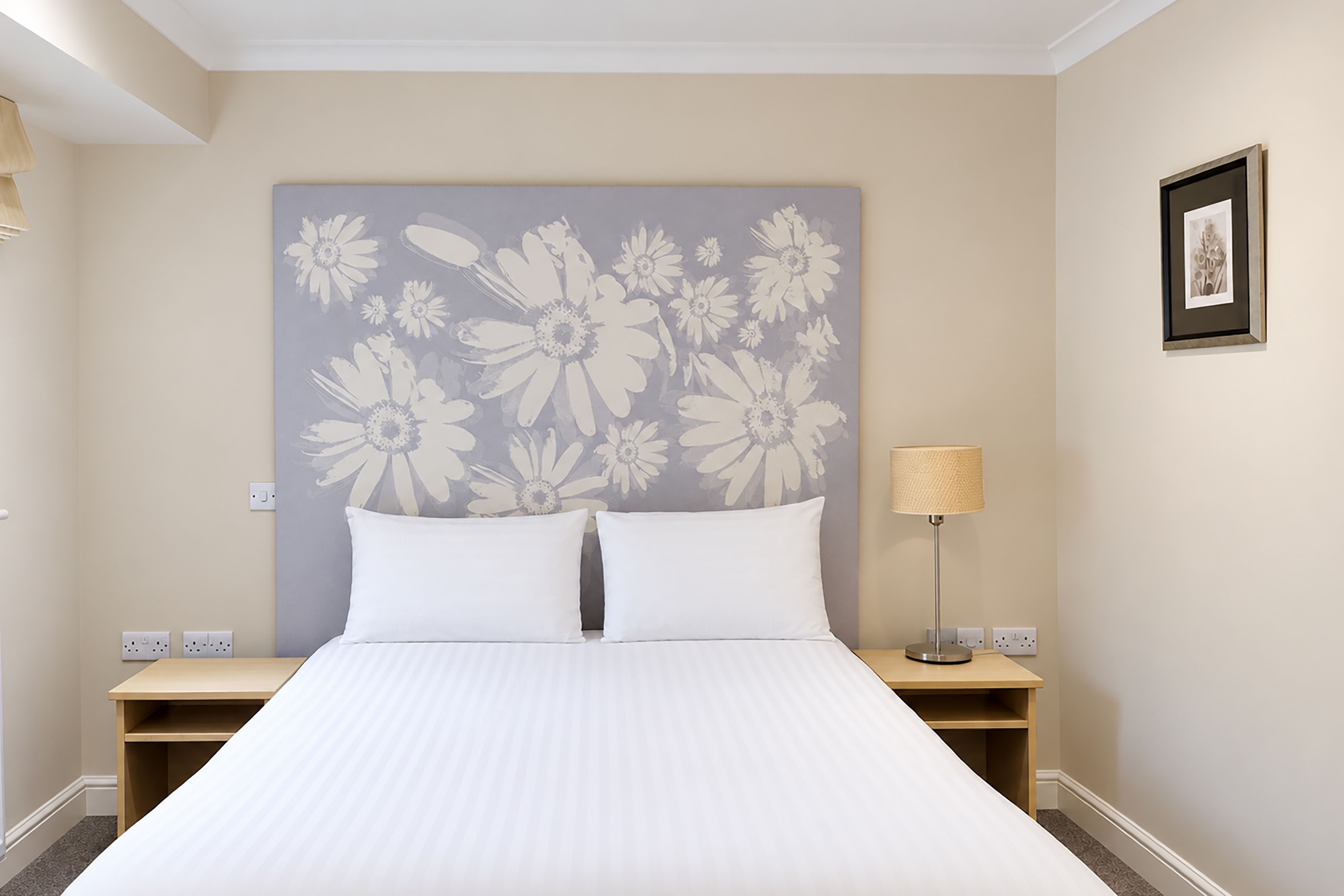 Une chambre d’hôtel avec un grand lit, deux tables de chevet, une lampe fixée au mur et un tableau encadré accroché au mur.