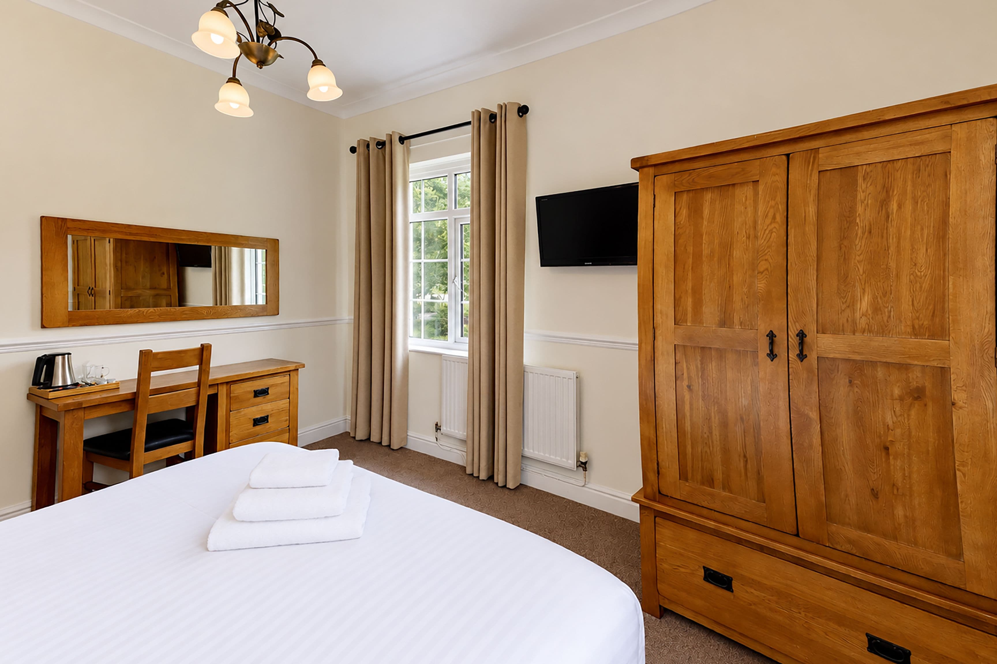Habitación de hotel con cama, escritorio de madera, silla, armario grande, televisor de pantalla plana y ventana con cortinas.