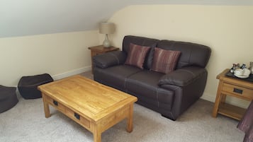 Una sala de estar con un sofá de cuero marrón oscuro, dos mesitas de madera, una mesa de centro de madera y una lámpara sobre una de las mesas.