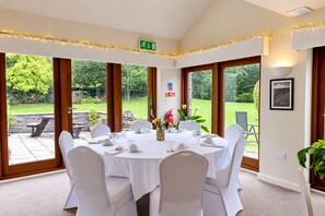 A dining area with a round table set for six, surrounded by white chairs, with a view of a garden through large glass doors. - Nant Ddu Lodge Hotel & Spa by Belvilla (Merthyr Tydfil)
