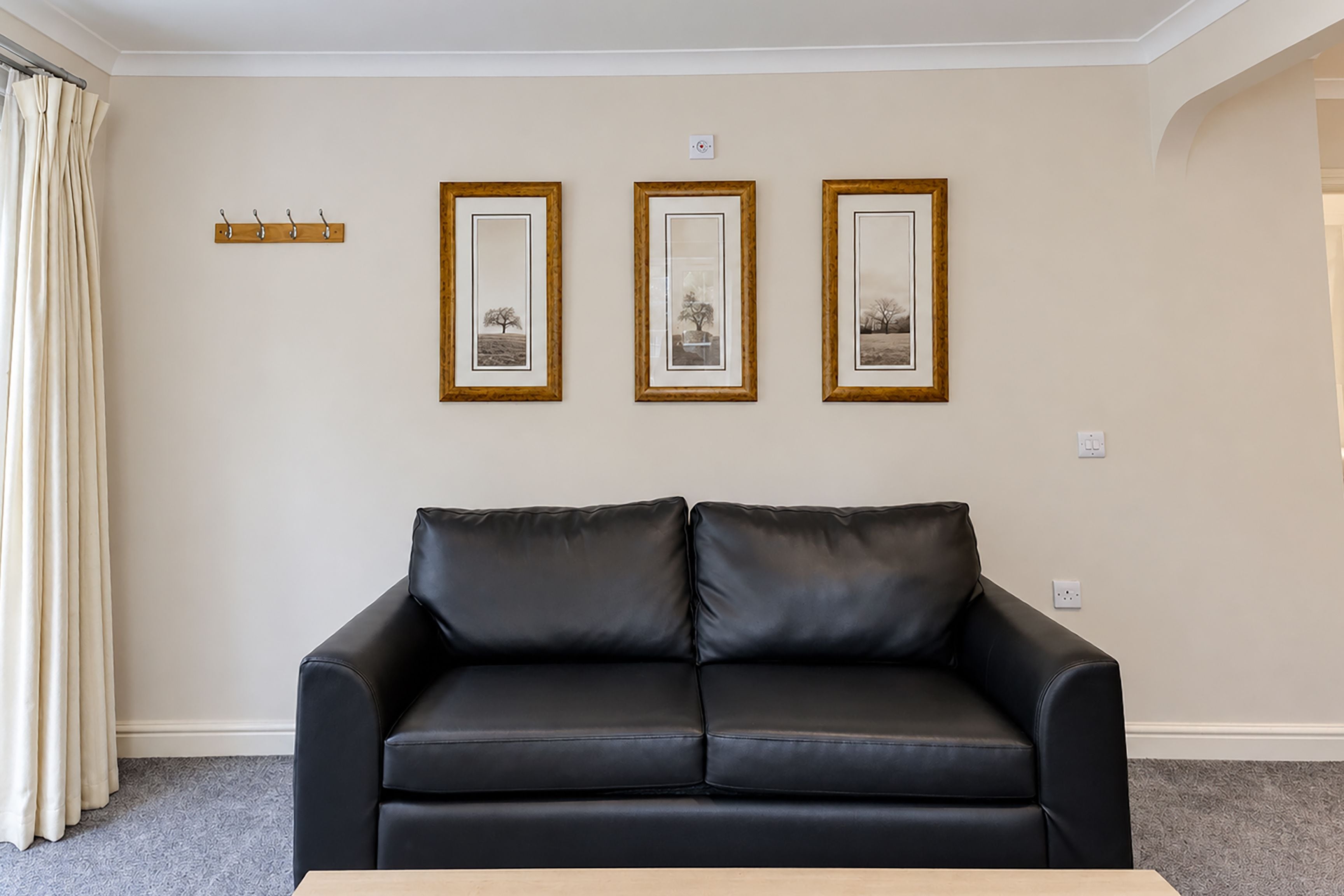 A black leather sofa in a living room with a light-colored carpet, three framed pictures on the wall, and a coat rack with hooks.