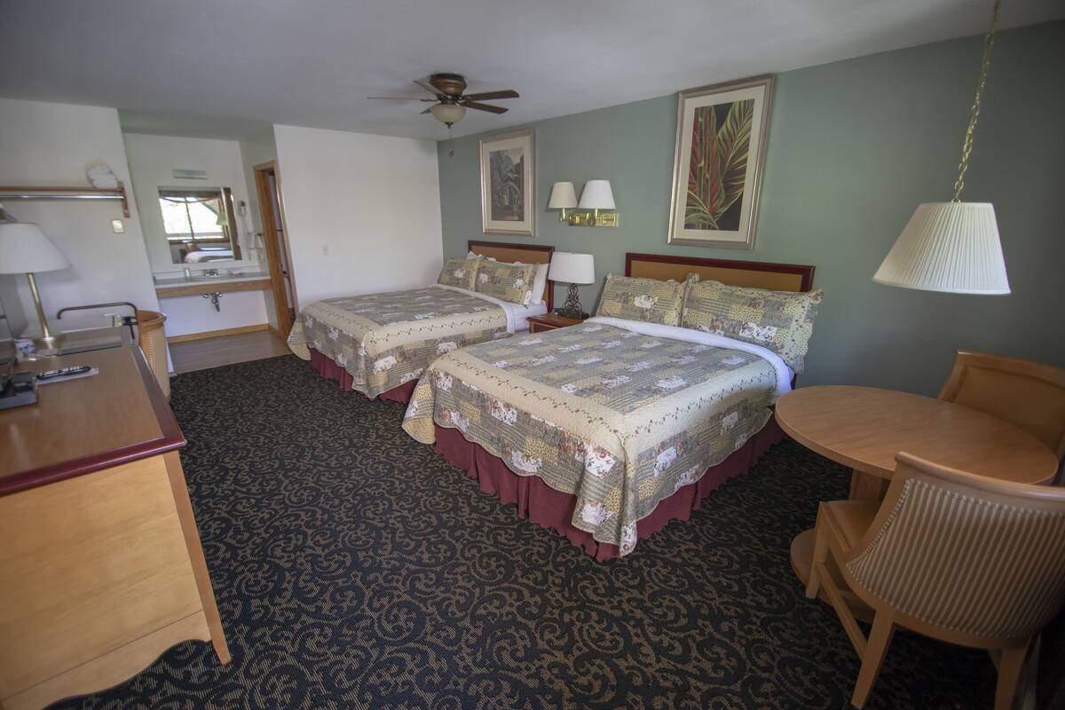 A hotel room with two beds, a desk, a chair, a table, a ceiling fan, and a lamp.