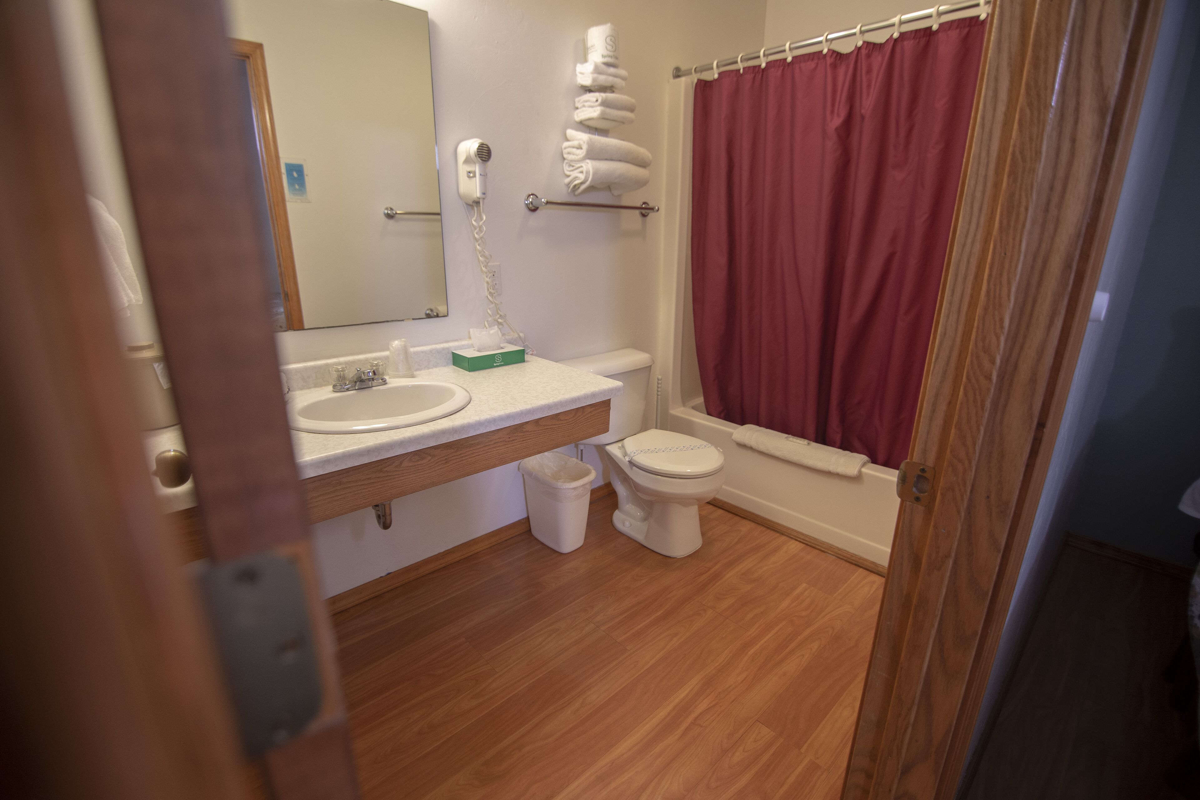 A bathroom with a wooden floor, white sink, toilet, and a red shower curtain.