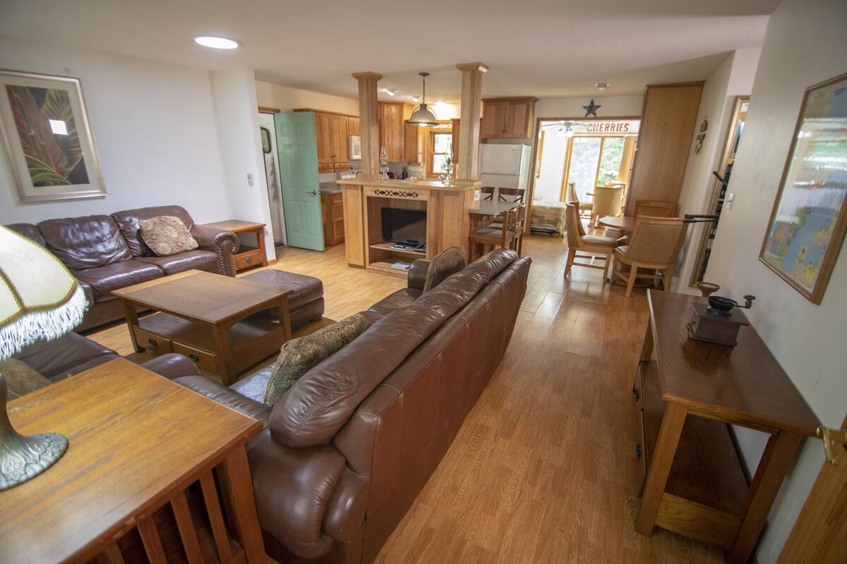 A spacious living area with a brown leather sofa, wooden coffee table, and a dining table with chairs. A kitchen with wooden cabinets and a central island is visible in the background.