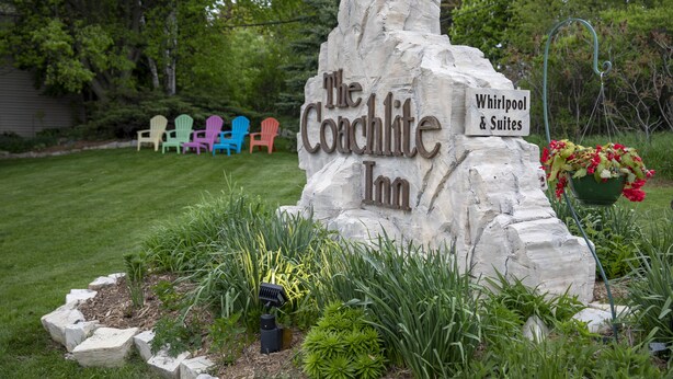 A sign for 'The Coachlite Inn' with a garden in front, including a stone planter with flowers and a lawn with colorful Adirondack chairs.