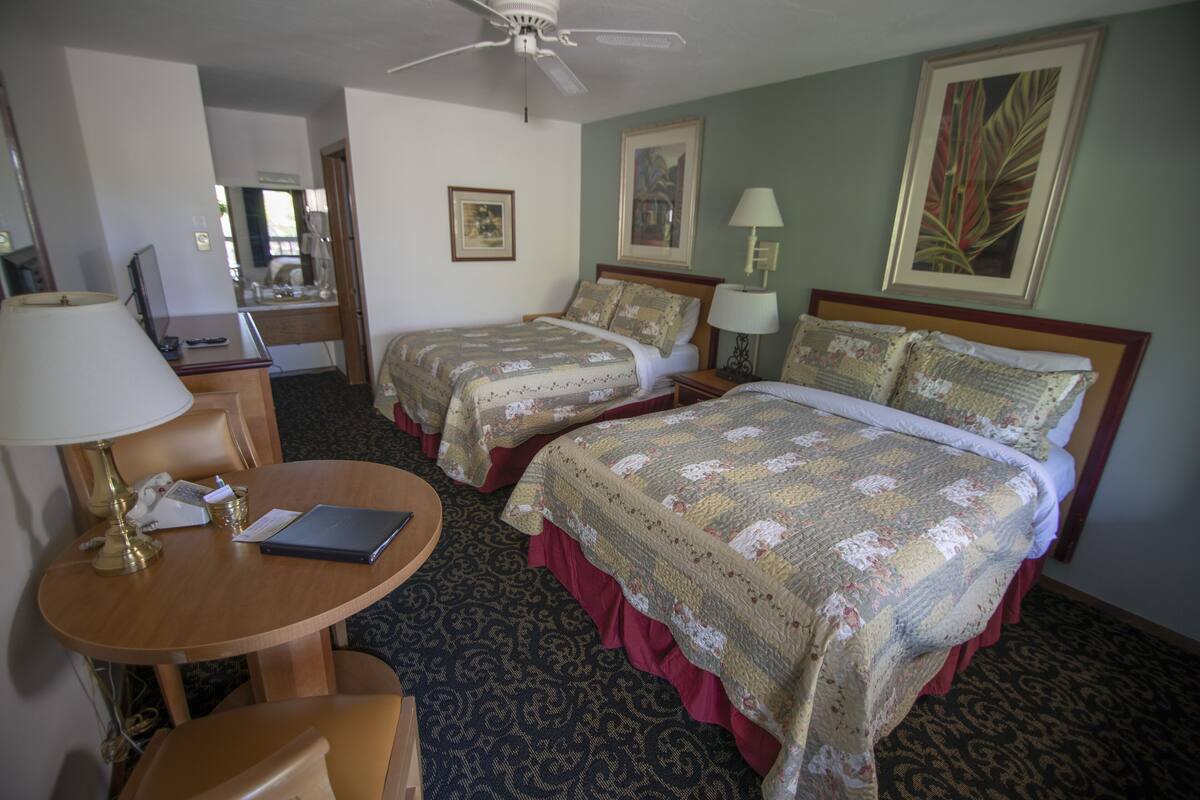 A hotel room with two beds, a desk with a lamp, a chair, and a ceiling fan.