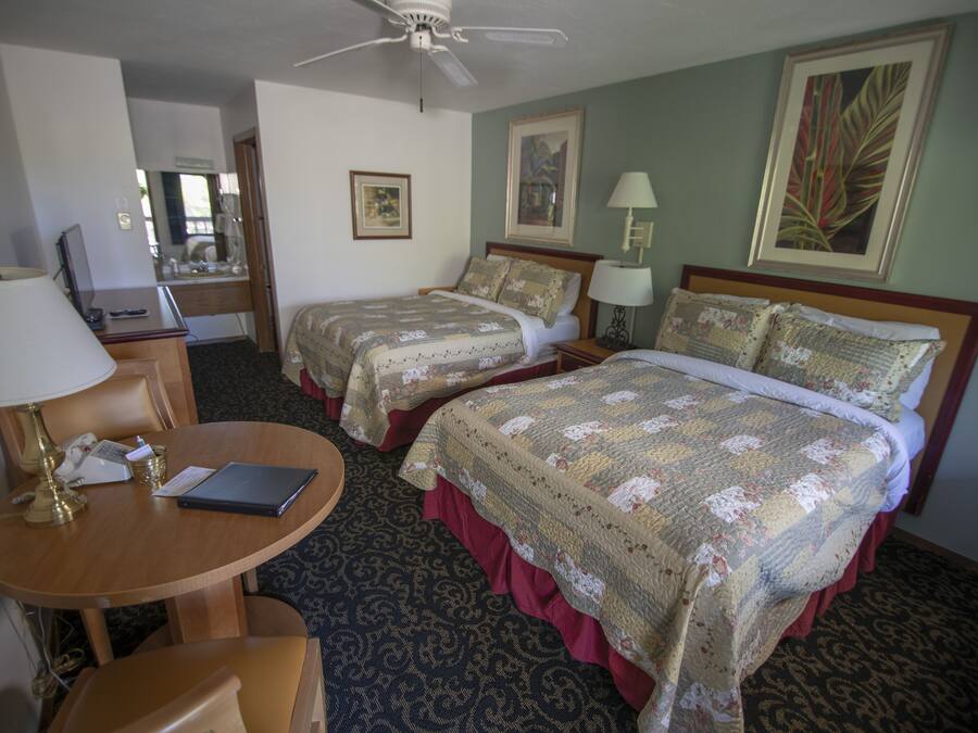A hotel room with two beds, a desk with a lamp, a chair, and a ceiling fan.