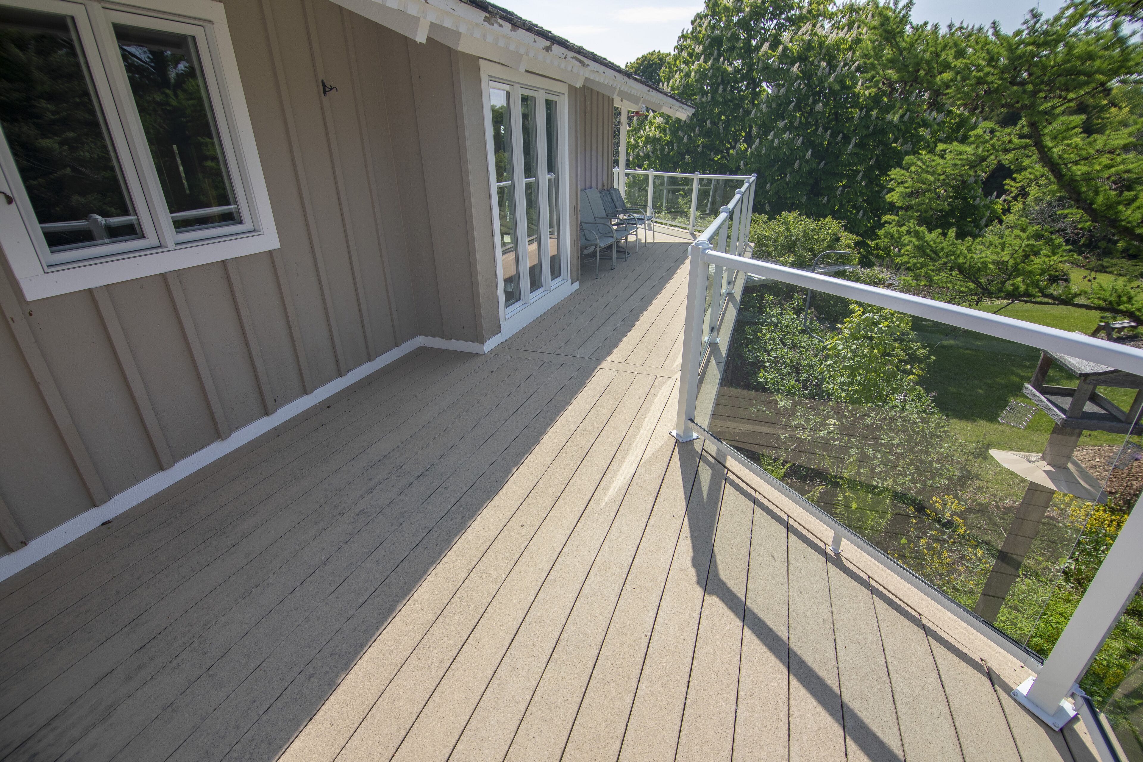 A wooden deck with a glass balustrade, a sliding glass door, and a seating area with two chairs.