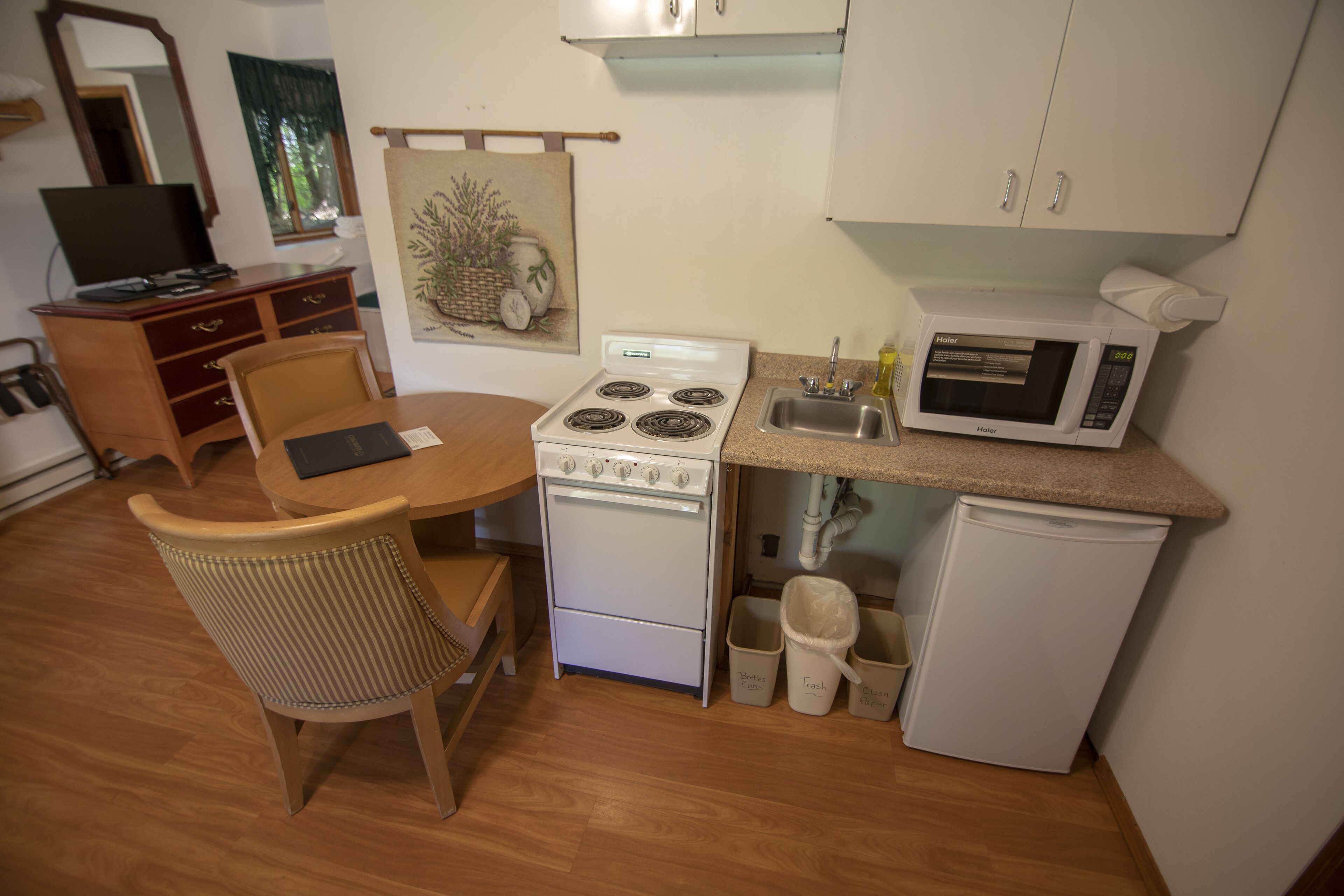 A compact kitchen with a stove, microwave, sink, and a small refrigerator. There is a dining table with chairs, a wooden cabinet, and a painting on the wall.