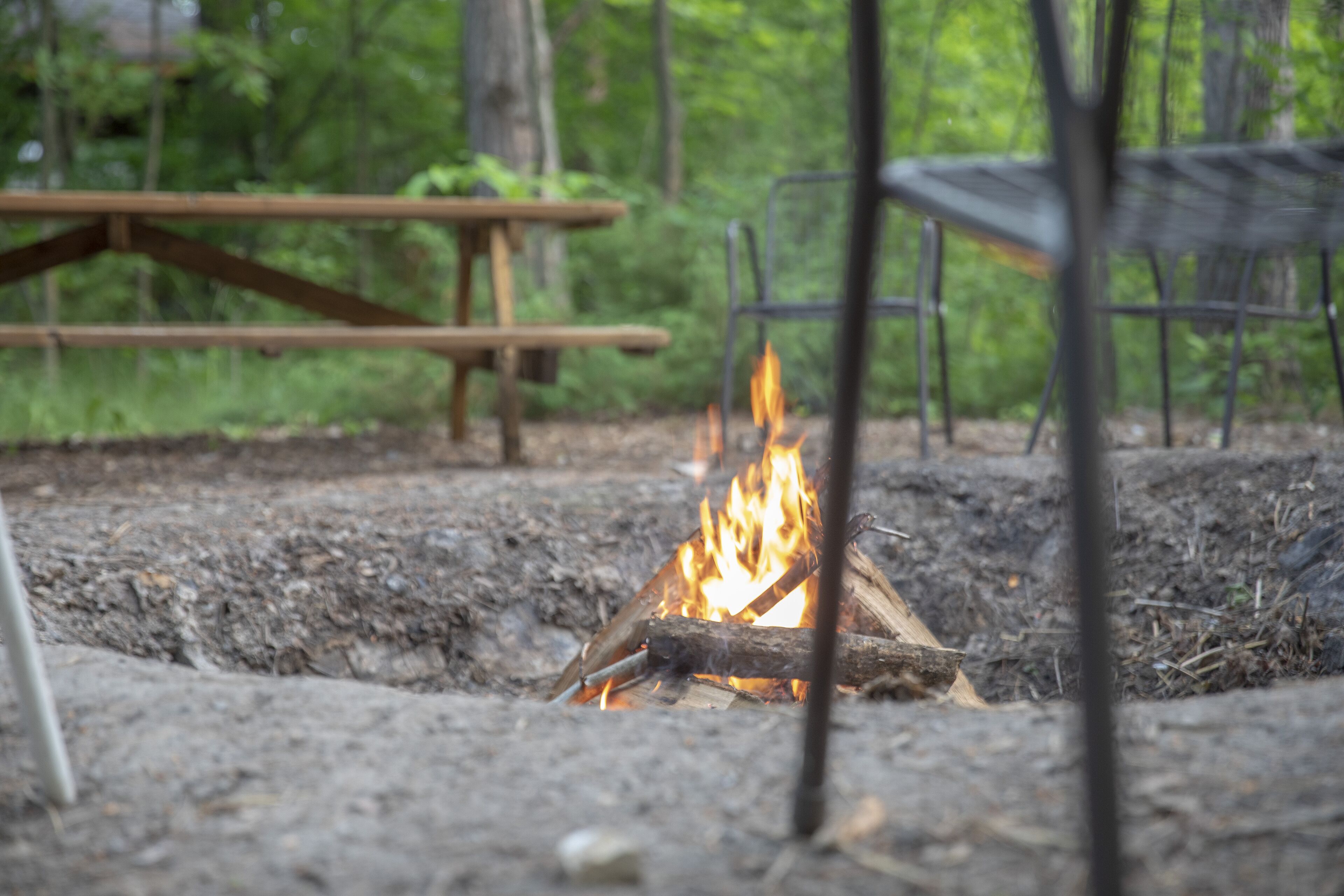 A campfire with a picnic table and metal chairs in a forested area.