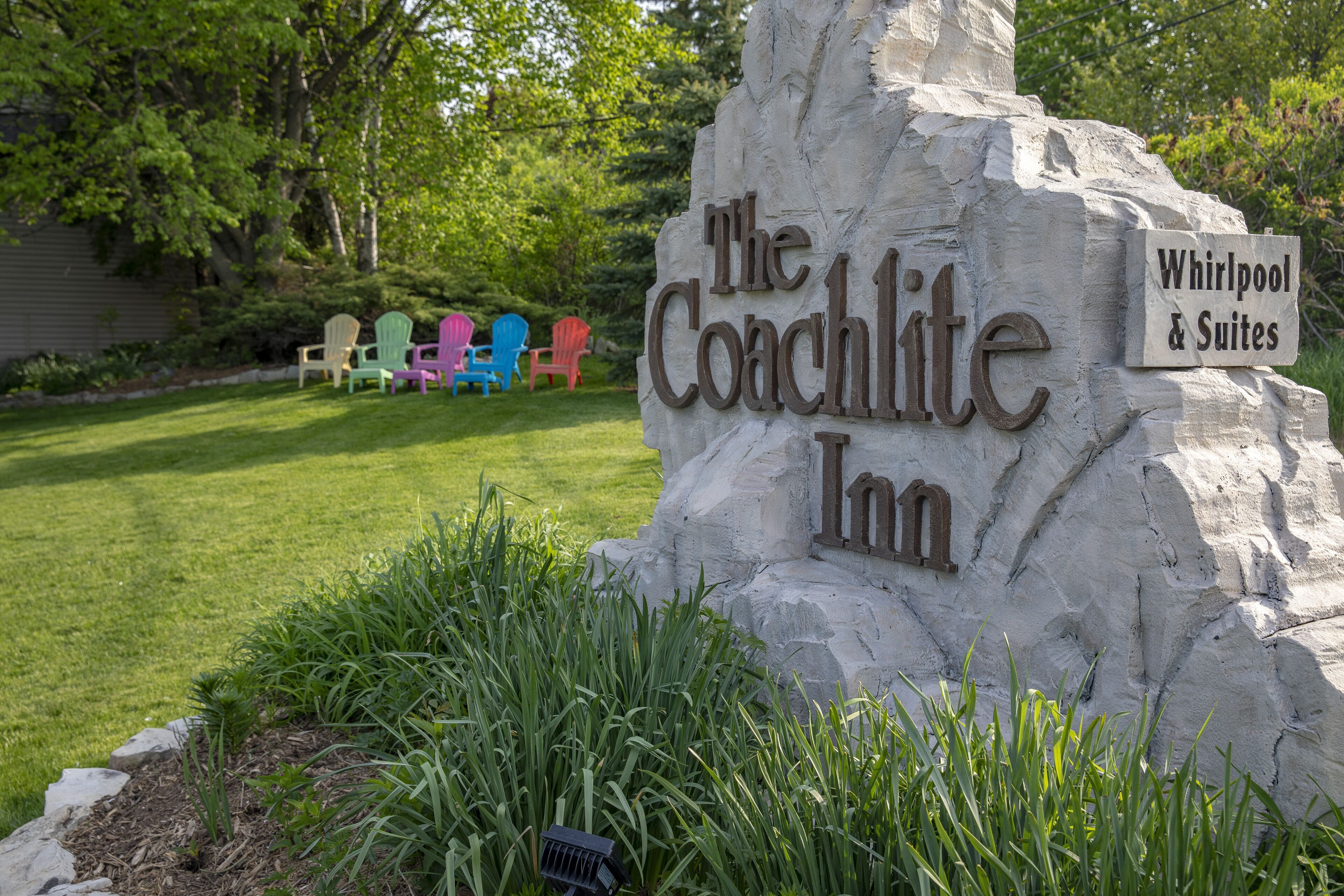 Une enseigne en pierre portant les inscriptions « The Coachlite Inn » et « Whirlpool & Suites », entourée d’une pelouse bien entretenue et de chaises Adirondack aux couleurs vives.