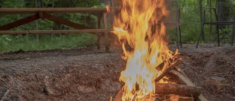 A campfire with a wooden bench and metal chairs in a forested area.